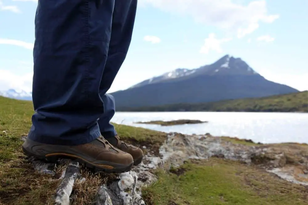 man wearing jeans and hiking boots standing near the lake and mountain