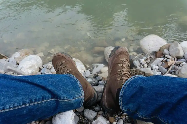 man wearing jeans and dark hiking boots sitting near a lake