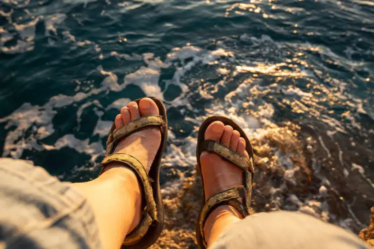 man wears open toe hiking sandals sitting near a lake