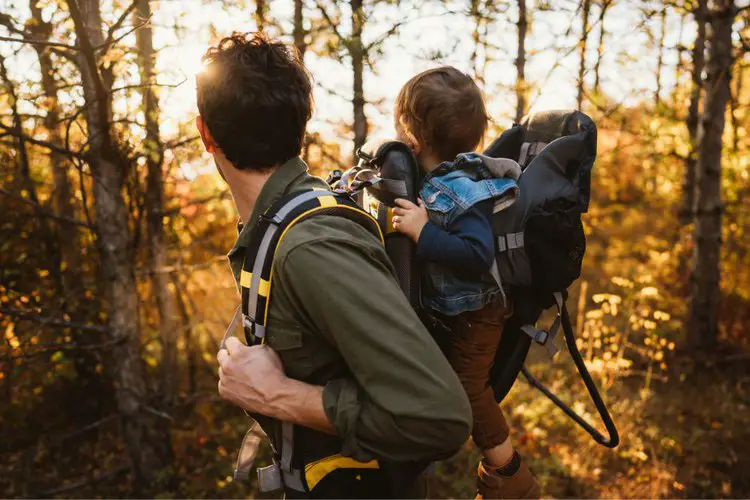 a man carries his baby in hiking backpack through a forest