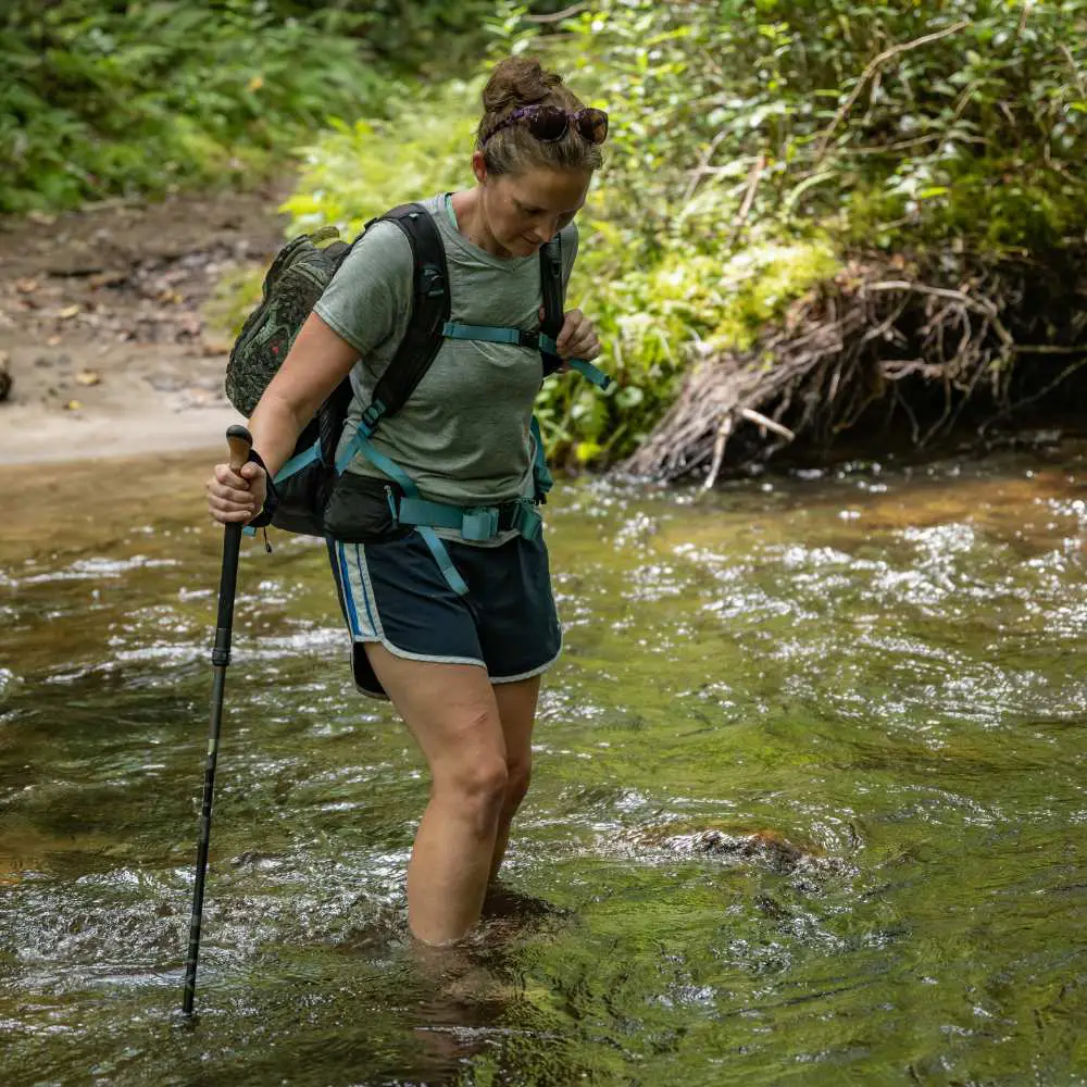 a woman is crossing the deep stream with a hiking pole