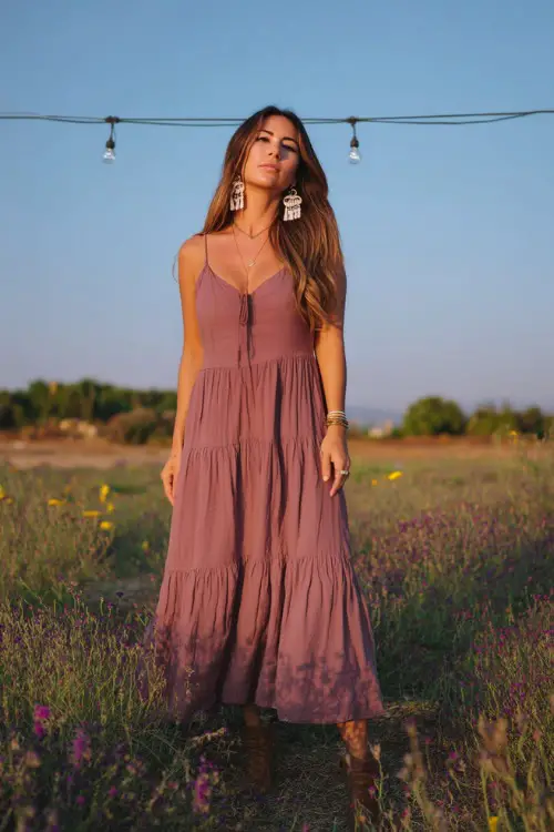 A confident woman in a sleeveless mauve flowy maxi dress with a tiered hem and boho earrings, standing in a field decorated with wildflowers and string lights 