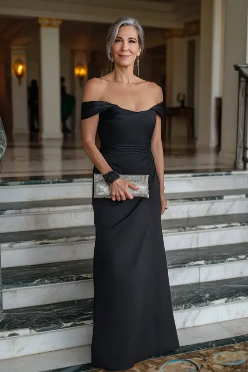 A confident woman over 50 in a black off-the-shoulder formal gown with a satin finish and silver clutch, posing on marble steps at a luxurious wedding hall entrance 