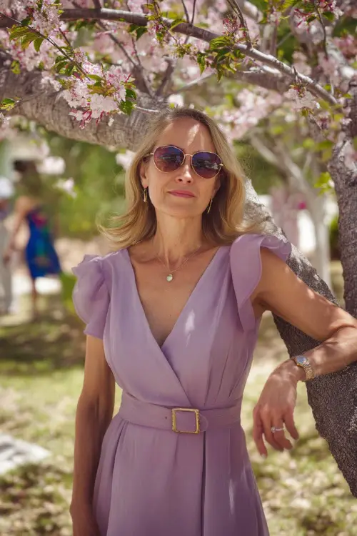 A confident woman over 50 in a knee-length lavender dress with a fitted waist and soft ruffle sleeves, standing under a blooming cherry blossom tree at a spring wedding 