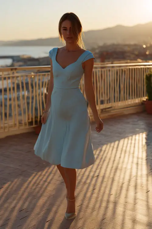 A fashionable woman in a powder blue cocktail dress with cap sleeves and a pleated skirt, walking across a rooftop terrace overlooking a coastal sunset