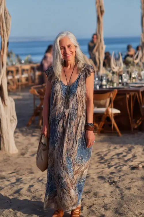 A fashionable woman over 50 in a flowy boho-style maxi dress with cap sleeves and woven sandals, standing at a beachside wedding setup with driftwood decor 