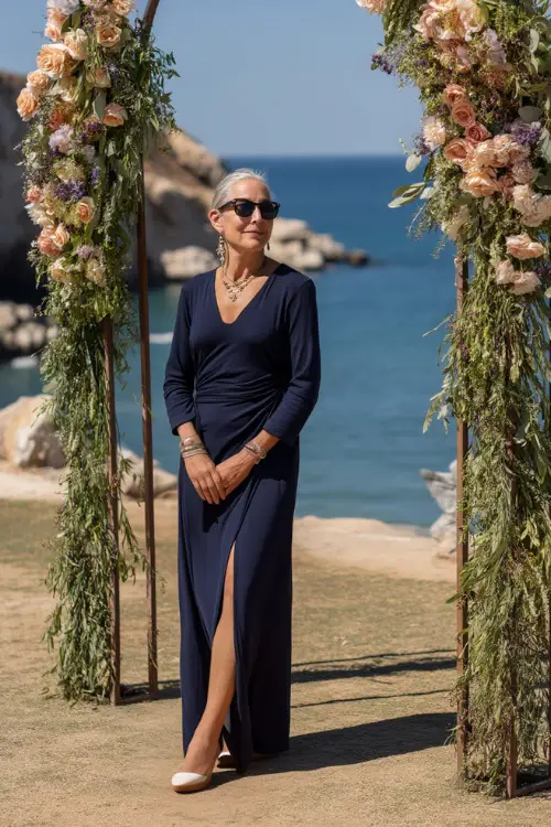 A fashionable woman over 50 in a navy blue long-sleeve maxi dress with a slit, gold accessories, and wedges, standing near a floral arch at a coastal wedding 