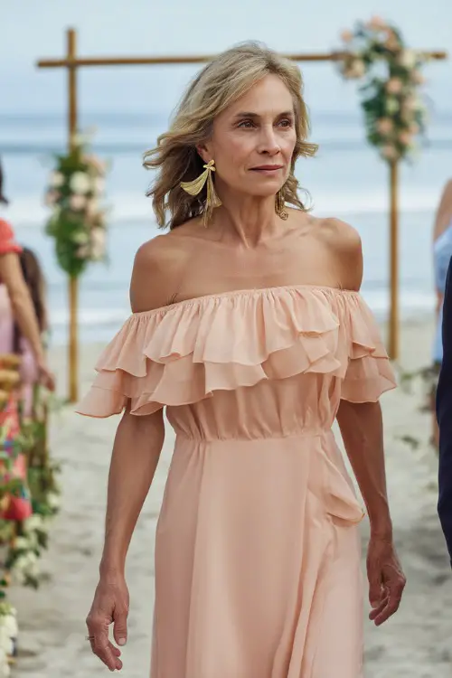 A graceful woman over 50 in a pastel peach off-the-shoulder maxi dress with ruffle trim and gold earrings, walking along a beachfront wedding aisle lined with simple floral decor