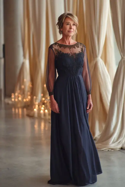 A graceful woman over 50 wearing a floor-length navy blue evening gown with sheer long sleeves and a beaded neckline, standing in a candlelit indoor wedding venue with elegant drapes 