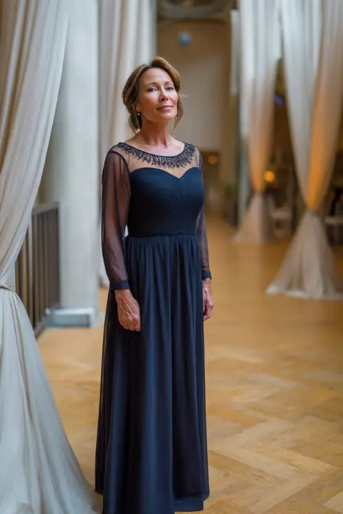 A graceful woman over 50 wearing a floor-length navy blue evening gown with sheer long sleeves and a beaded neckline, standing in a candlelit indoor wedding venue with elegant drapes