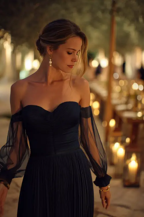 A graceful woman wearing a navy blue pleated gown with sheer flutter sleeves and pearl earrings, walking through a candlelit outdoor reception area 