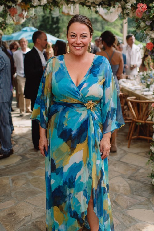 A plus-size woman over 40 wears a flowing wrap maxi dress in warm cobalt blue chiffon with a bold watercolour brushstroke print in navy, gold, and jade, a deep V-neckline