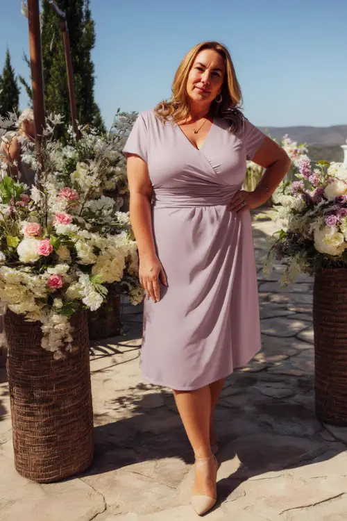 A plus-size woman over 50 wearing a soft lavender wrap dress with short sleeves and neutral heels, standing beside floral arrangements at an outdoor spring wedding