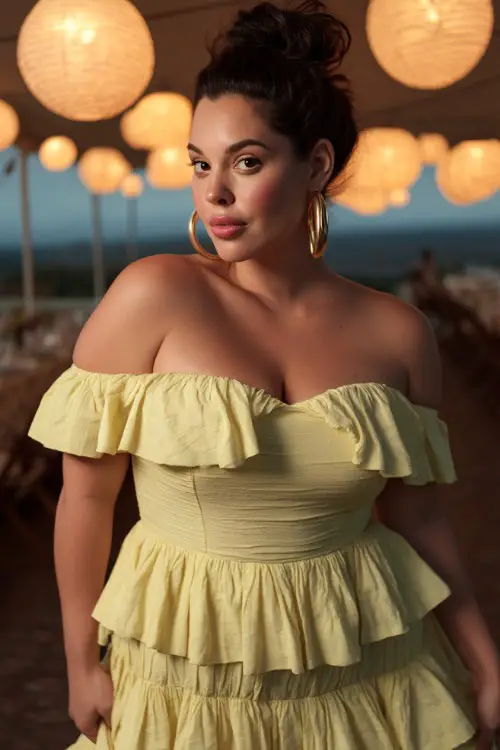 A plus-size woman wearing a pastel yellow off-the-shoulder dress with tiered ruffles and gold hoop earrings, posing under lanterns at an open-air evening reception