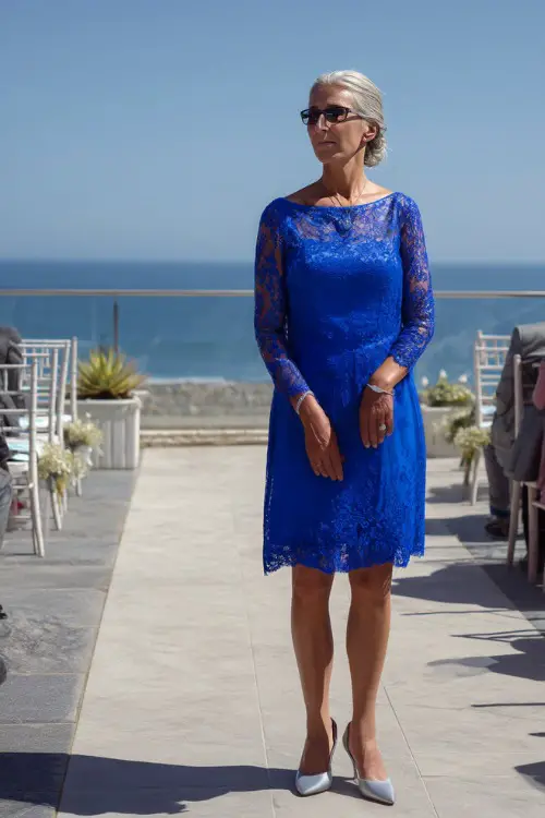 A poised woman over 50 in a royal blue A-line dress with long lace sleeves and silver heels, standing on a terrace overlooking a seaside wedding ceremony 