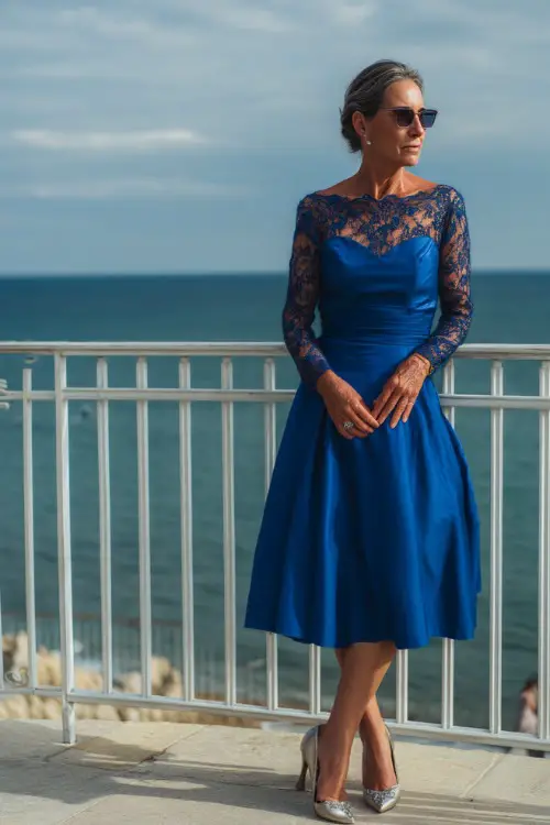 A poised woman over 50 in a royal blue A-line dress with long lace sleeves and silver heels, standing on a terrace overlooking a seaside wedding ceremony