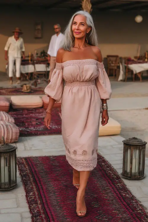 A poised woman over 50 wearing a blush pink off-the-shoulder midi dress with delicate floral embroidery and heels, walking near a minimalist boho-style wedding setup with rugs and lanterns