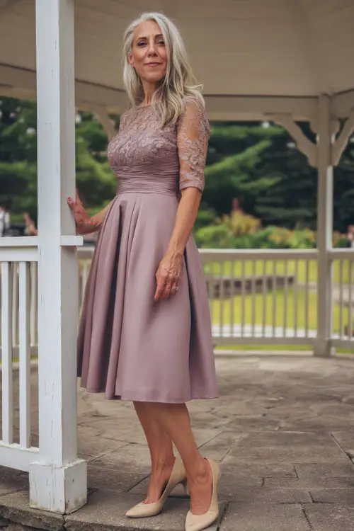 A sophisticated woman over 50 wearing a tea-length mauve A-line dress with lace sleeves and beige pumps, posing under a white gazebo at a garden wedding