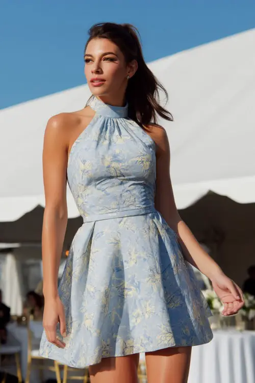 A stylish woman in a light blue floral-print cocktail dress with a high neckline and short hem, standing in front of a white tent at an elegant outdoor wedding reception