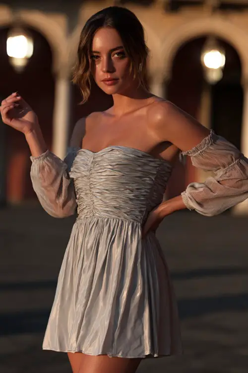 A stylish woman in a pale silver cocktail dress with a ruched bodice and chiffon sleeves, walking across a courtyard lit by lanterns at a sunset ceremony 