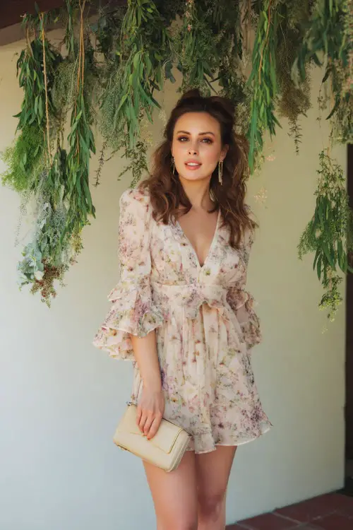 A stylish woman in a soft pastel floral mini dress with ruffled sleeves and a matching clutch, posing beside a hanging greenery installation at a boho-chic wedding 