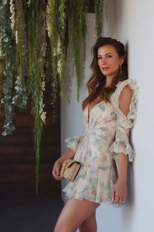 A stylish woman in a soft pastel floral mini dress with ruffled sleeves and a matching clutch, posing beside a hanging greenery installation at a boho-chic wedding 