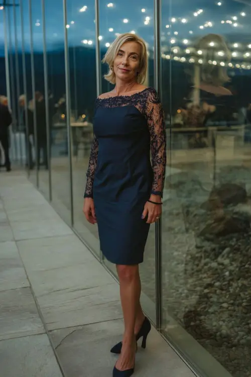 A stylish woman over 50 in a knee-length navy blue cocktail dress with lace sleeves and pointed heels, standing in front of a modern wedding venue with glass walls and evening lights 