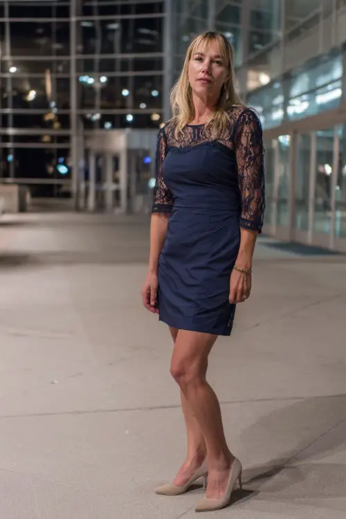A stylish woman over 50 in a knee-length navy blue cocktail dress with lace sleeves and pointed heels, standing in front of a modern wedding venue with glass walls and evening lights 