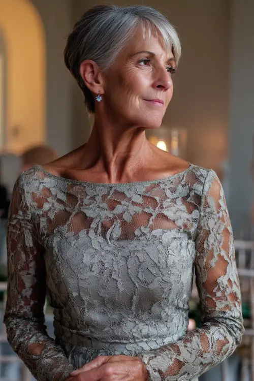 A stylish woman over 50 in a midi-length silver lace dress with a fitted silhouette and sparkly earrings, standing under soft lighting in an indoor wedding venue