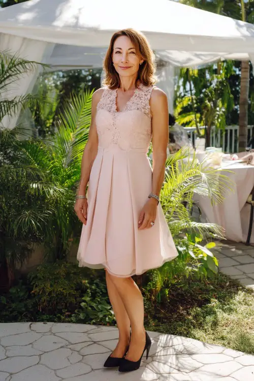 A stylish woman over 50 in a sleeveless blush pink fit-and-flare dress with lace detailing and flats, standing under a white tent surrounded by greenery for a garden wedding in full sunlight 