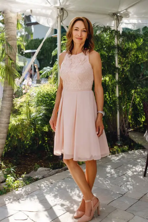 A stylish woman over 50 in a sleeveless blush pink fit-and-flare dress with lace detailing and flats, standing under a white tent surrounded by greenery for a garden wedding in full sunlight 