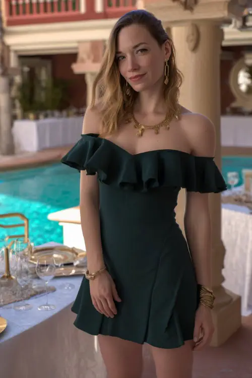 A woman in a dark green cocktail dress with short ruffled sleeves and gold jewelry, standing beside a poolside reception area set for an elegant evening