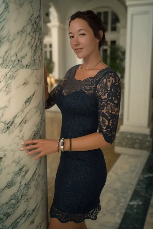 A woman in a dark navy lace cocktail dress with scalloped hem and three-quarter sleeves, posing beside a marble column at a luxury summer wedding reception 