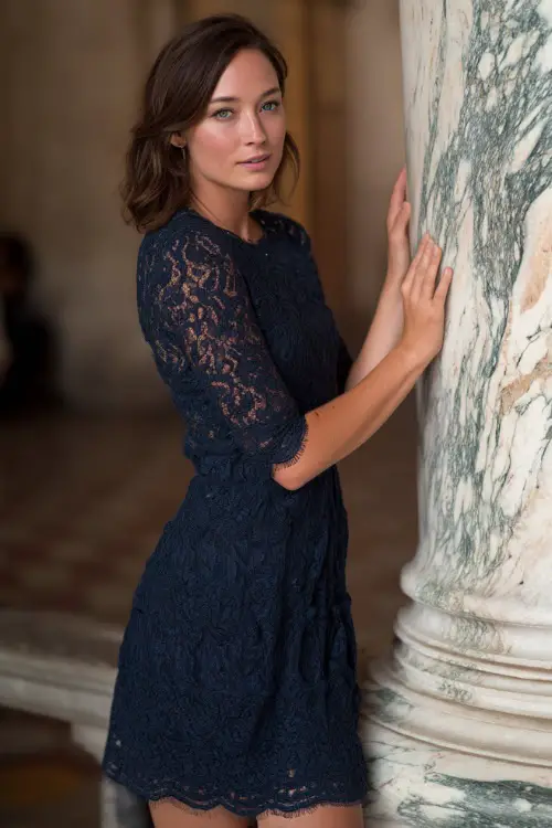 A woman in a dark navy lace cocktail dress with scalloped hem and three-quarter sleeves, posing beside a marble column at a luxury summer wedding reception