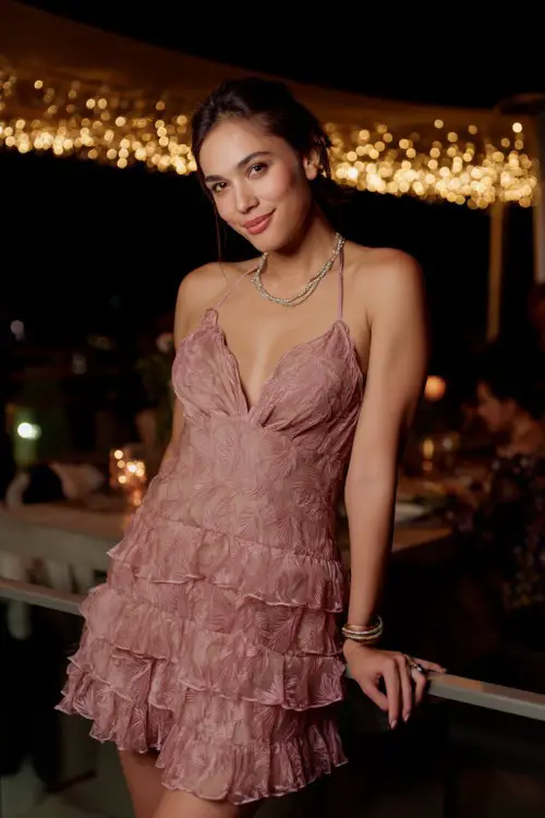 A woman in a fitted dusty rose floral cocktail dress with ruffle hem and delicate jewelry, posing at a rooftop wedding reception decorated with twinkle lights 