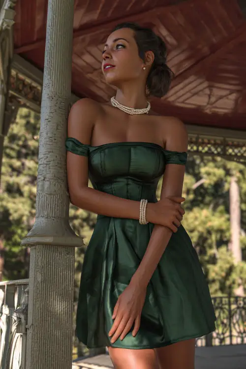A woman in a forest green off-the-shoulder cocktail dress with a structured bodice and pearl accessories, standing beneath an ornate gazebo at a luxury summer venue