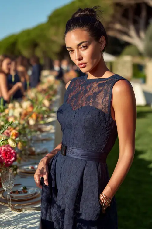 A woman in a sleeveless navy blue tea-length dress with lace overlay and a structured waist, standing near a long outdoor reception table lined with florals
