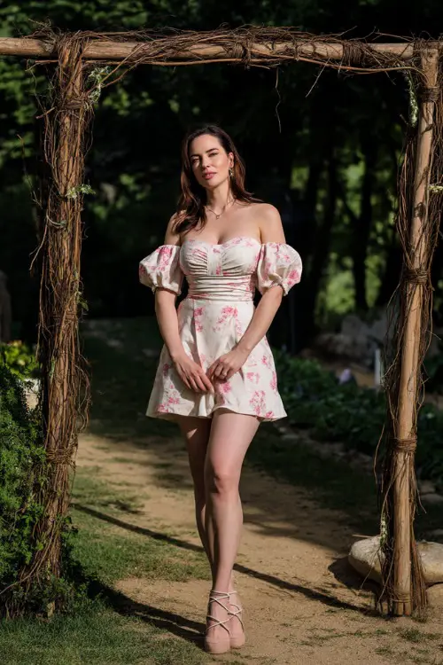 A woman in a white and pink floral cocktail dress with puff sleeves and strappy sandals, standing beside a rustic wooden arch at a garden wedding