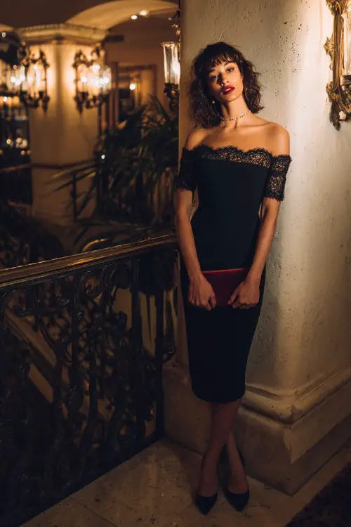 A woman over 30 wears a black off-shoulder dress with intricate lace embroidery along the neckline and sleeves, styled with sleek pumps and a velvet clutch