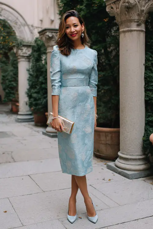 A woman over 30 wears a soft blue jacquard dress with subtle floral patterns woven into the fabric, styled with pointed-toe pumps