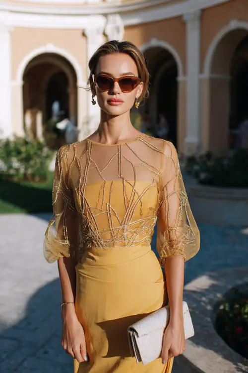 A woman over 40 wears a burnished gold dress with sheer sleeves detailed with abstract geometric embroidery and fluid draping through the skirt