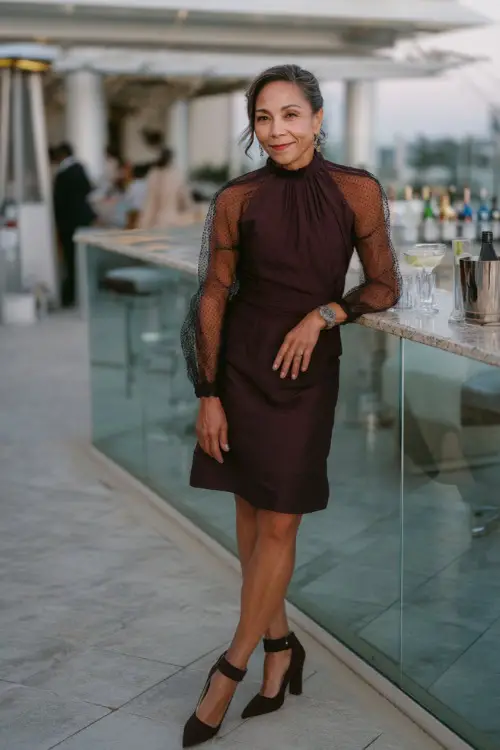 A woman over 50 in a dark plum cocktail dress with a high neckline, sheer sleeves, and block heels, posing near a bar setup at a rooftop wedding reception 