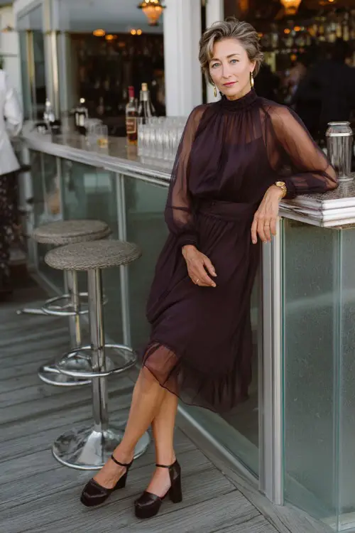 A woman. over 50 in a dark plum cocktail dress with a high neckline, sheer sleeves, and block heels, posing near a bar setup at a rooftop wedding reception