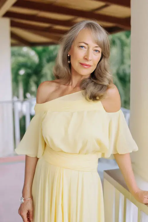 A woman over 50 in a pastel yellow chiffon dress with flutter sleeves and a sash waist, posing on a terrace at a warm-weather destination wedding