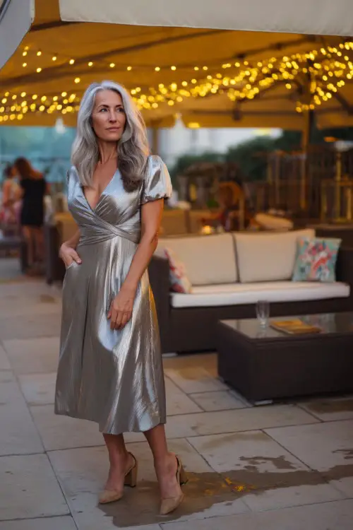 A woman over 50 in a short-sleeve metallic silver midi dress with block heels, standing beside an outdoor lounge area under a canopy of fairy lights 