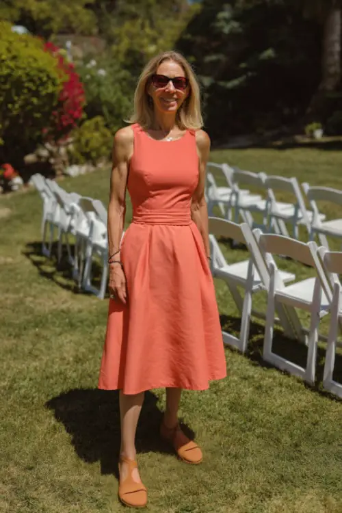 A woman over 50 in a sleeveless coral midi dress with a flared skirt and tan sandals, standing on a sunny lawn with white folding chairs arranged for a summer garden wedding 