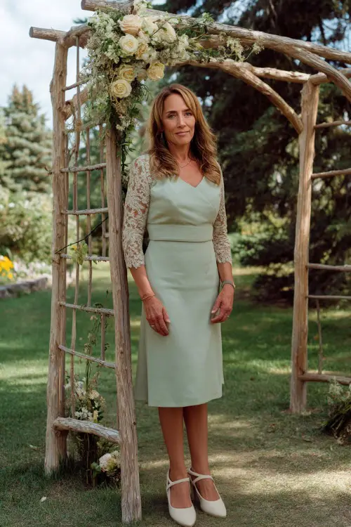 A woman over 50 in a soft sage green midi dress with lace sleeves and beige pumps, standing near a rustic wooden wedding arch with light spring florals 