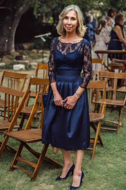 A woman over 50 wearing a midi-length navy blue dress with elbow-length lace sleeves and pumps, standing near wooden chairs at a classic outdoor wedding