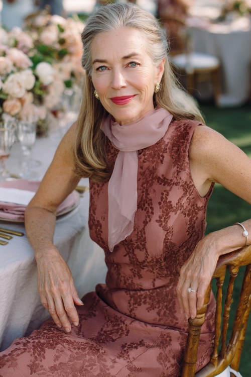 A woman over 50 wears a sleeveless sheath dress in rich dusty rose crepe with an all-over tonal burnout velvet pattern of climbing wisteria blooms, a graceful bateau neckline, and a scalloped heml