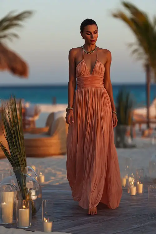 A classy woman in a pale coral formal dress with thin straps and a pleated skirt, walking beside floating candles and palm decor at a sunset beachfront reception 
